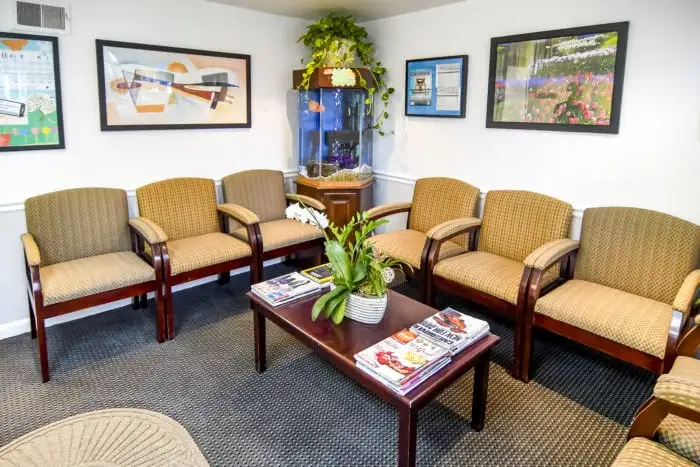 Comfortable waiting room with beige armchairs arranged around a dark wooden coffee table, framed artwork on the walls, and a decorative plant in the corner.