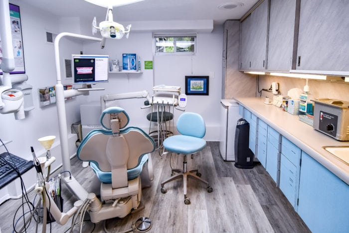 Interior view of a modern dental treatment room featuring a beige patient chair with blue accents, light blue cabinetry with light wood countertops along the right wall, overhead dental lighting, wall-mounted monitors, and a rolling stool on light gray wood-look flooring.