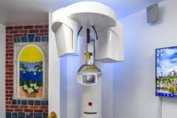 White Cone Beam CT scanner standing in a treatment room with a brick wall and decorative window in the background.
