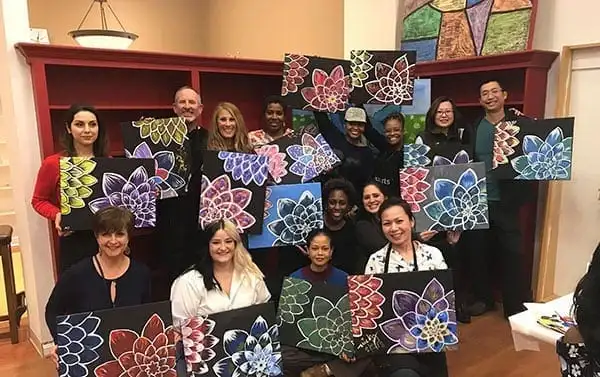 Dental office team members posing together while holding up colorful abstract artwork, standing in front of wooden cabinetry in the office.